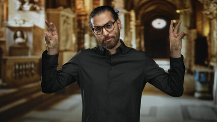Obraz premium Man with beard and glasses, hands raised in prayerlike gesture wearing black shirt inside a building aisle with ornate columns; calm reflection.