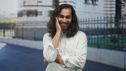 Man smiling with hand on cheek and folded arm standing on street in front of building and iron fence, wearing white shirt; quiet gentle contentment reflection.