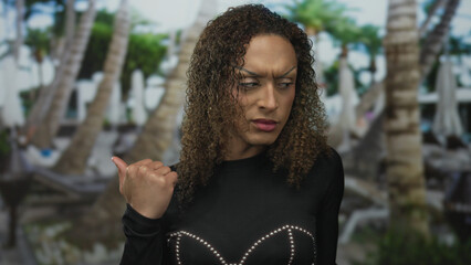 Transgender woman frowning and pointing her thumb at a pool by a resort building outdoors under...