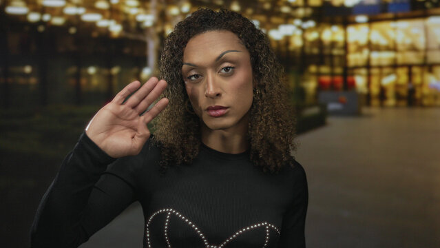 Transgender woman in black studded top waves hand on street under glowing streetlights at dusk; pride confidence visibility.