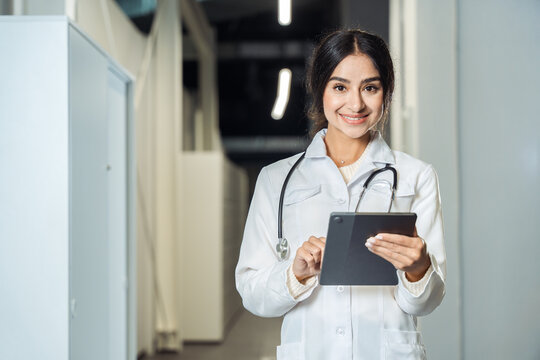 Smiling female doctor standing in hospital corridor and using digital tablet. Modern healthcare, medicine and technology concept.
