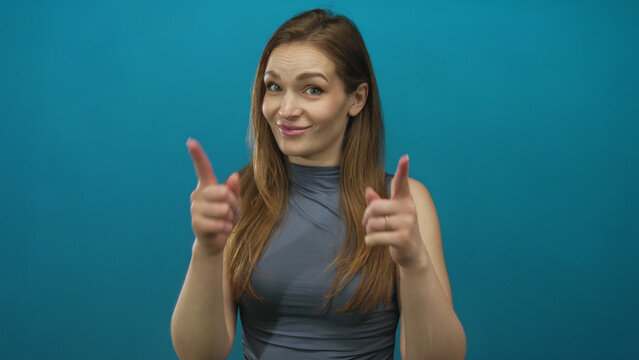 Young caucasian woman finger pointing with both hands in blue studio setting while smiling confidently; playfulness motivation encouragement positivity.