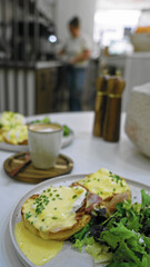 Table with eggs benedict and salad in a modern cafeteria with a woman seen in the background near kitchen area blurry and out of focus.