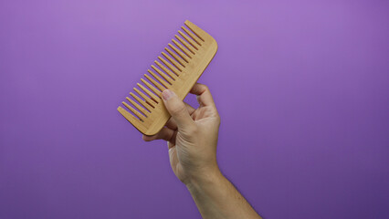 Naklejka premium Male hand holding wooden comb against purple background showcasing simplicity and isolation in design concept.
