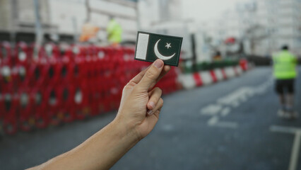 Man holding a small embroidered pakistan flag in city street environment outdoors with traffic barriers in background.