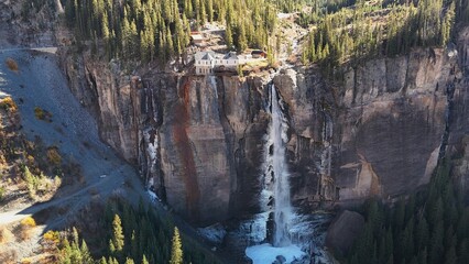 Aerial view of Bridal Veil Falls cascading down a steep cliff face, with a historic structure perched precariously above, amidst the golden hues of autumn foliage, Telluride, Colorado, United States.