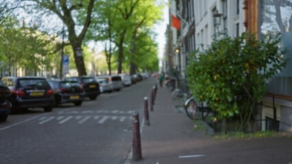 Blurred view of an amsterdam street showcasing parked cars, dutch license plates, lush green trees, and traditional architecture under bright daylight conditions.