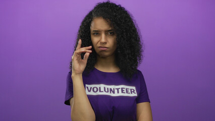 Young hispanic woman wearing a volunteer shirt stands against an isolated purple background, appearing thoughtful and expressive.
