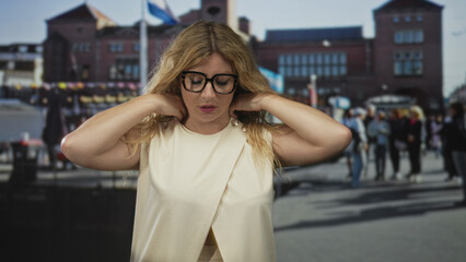 Fototapeta premium Woman adjusts neckline and touches neck wearing glasses on city street in front of brick building; contemplation.