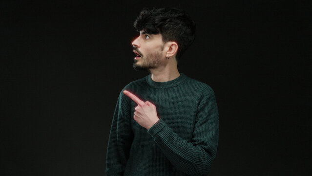 Young man in green sweater with surprised expression points to side against dark background conveying curiosity and intrigue.