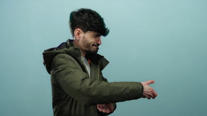 Young man pulling imaginary rope against a blue background, wearing a green jacket and looking...
