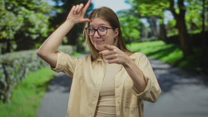 Woman in beige shirt frames l with fingers on forehead in green forest path near stone wall; playful teasing.