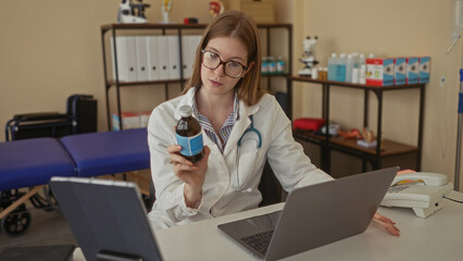 Young blonde woman doctor in white coat holds amber medicine bottle while browsing on laptop in...