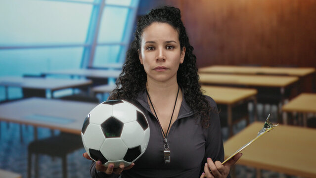 Hispanic woman holding soccer ball and clipboard in classroom with focused expression, teaching sports indoors in educational setting, wearing whistle and dark jacket - Powered by Adobe