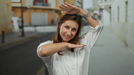 Woman making frame gesture with hands in city street, smiling in casual white shirt with tattoo visible, young and relaxed, outdoors during daylight.