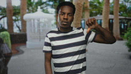 Fototapeta premium Young african american man wearing striped shirt making thumbs down gesture on a street near trees; disapproval.