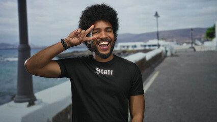 African american man in staff uniform makes victory gesture outdoors on seaside promenade, smiling brightly under cloudy skies with serene sea view.
