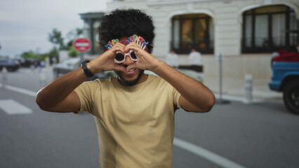 Man with birthday glasses smiling in a city street, making heart gesture, embodying joy and celebration in an urban outdoor setting.