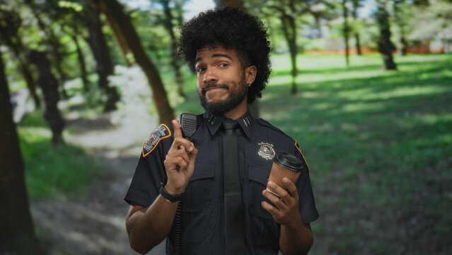 Police officer in uniform holds coffee cup and gestures in a sunny park setting surrounded by green trees. - Powered by Adobe