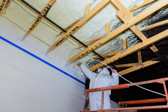 Painter in protective gear uses spray machine to apply refractory paint insulation to foam ceiling beams of construction site.