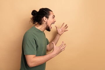 Excited young man with beard wearing a green t shirt shouts with open hands against beige...