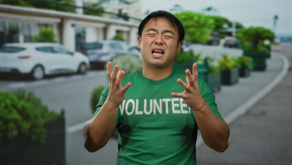 Naklejka premium Young man wearing green volunteer t-shirt gesturing emotionally on a busy city street with cars and greenery in an urban setting.