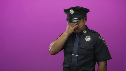 Young chinese policeman with visible hat and badge covers face with hand in purple studio; stress...