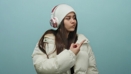 Young woman wearing headphones and white jacket pointing with a contemplative expression against a blue background, showcasing modern style and technology.