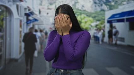 Fototapeta premium Woman covering face with hands on busy crowded street lined with shops; embarrassment selfconsciousness.