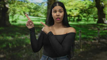 Woman with open mouth finger pointing upward in forest clearing under green canopy amid tall tree...