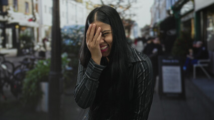 Obraz premium Latin woman with long dark hair covering her face with her hand on street while laughing in a candid moment; happiness.