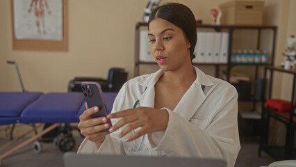 Woman doctor holds smartphone in clinic with stethoscope draped around neck and blue examination table visible behind her; telehealth empathy.