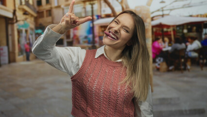 Woman smiling doing rock gesture on city terrace with people in background enjoying coffee at...