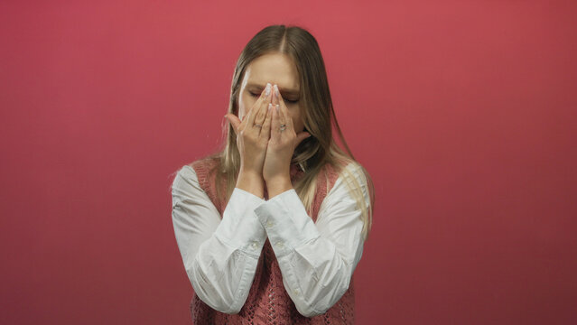 Woman expressing frustration holding her head against an isolated red background, wearing a white shirt and pink vest, showcasing emotion in a simple clean setting.