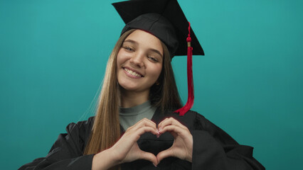 Girl wearing graduation cap makes heart gesture in studio with turquoise background; pride success...