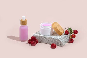Pink skincare serum and cream with raspberries on on a stone podium on a beige background.