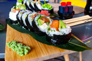Fresh sushi rolls served on a bamboo leaf over a wooden tray, accompanied by wasabi and soy sauce bottles in the background.
