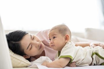 Loving asian mom carying of her newborn baby at home. Mother and baby boy playing in sunny bedroom. Parent and little kid relaxing at home. Family having fun together. Childcare, maternity concept.