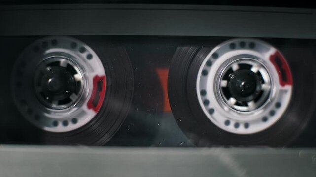 Close-up shot of a vintage audio cassette's transparent reels rotating during playback. The dark magnetic tape spools from left to right inside the classic analog media format