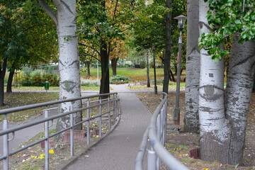 Paved pedestrian path with handrails leading through a green park with trees and lamps.