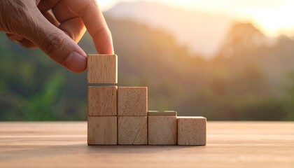 Hand places a block atop a growing structure made of wooden blocks, blurred nature background, warm light