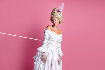 Fototapeta premium Gorgeous woman tied with rope in vintage baroque gown showcasing antique style against pink background
