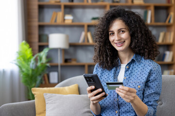 Young woman on sofa smiling while holding smartphone and credit card, making a secure online payment at home casual, relaxed mobile shopping and mobile banking experience