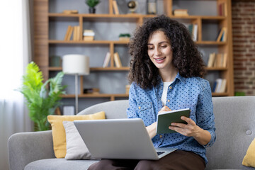 Young woman with curly hair confidently sitting on a sofa, taking notes in a notebook while using a laptop for remote work or online education in a cozy home environment