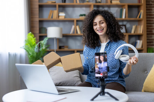 Young woman smiling, presenting new wireless headphones from an open cardboard box to a smartphone on a tripod, creating content for her social media vlog