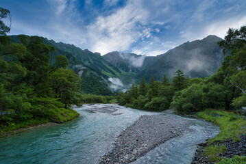 Serene river and lush green mountains in Kamikochi, Nagano, Japan. Morning mist rises over the clear blue water of the Azusa River surrounded by forest and peaks of the Northern Japan Alps, no people.