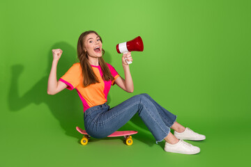 Young woman on skateboard with megaphone celebrating on bright green studio background wearing a bold orange and pink shirt and jeans