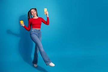 Energetic young woman holding a smartphone and cup on a vibrant blue background expressing a...