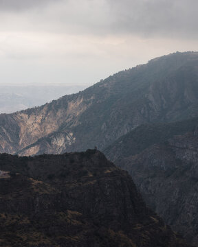 View of rugged, dark mountains reaching towards a cloudy sky, juxtaposed with the lighter tones of the distant landscape, Tatev, Syunik Province, Armenia.