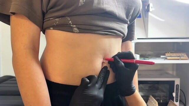 A young woman with light skin is getting a navel piercing. A tattoo artist marks the spot with a red pen. The setting is a clean, professional studio.
